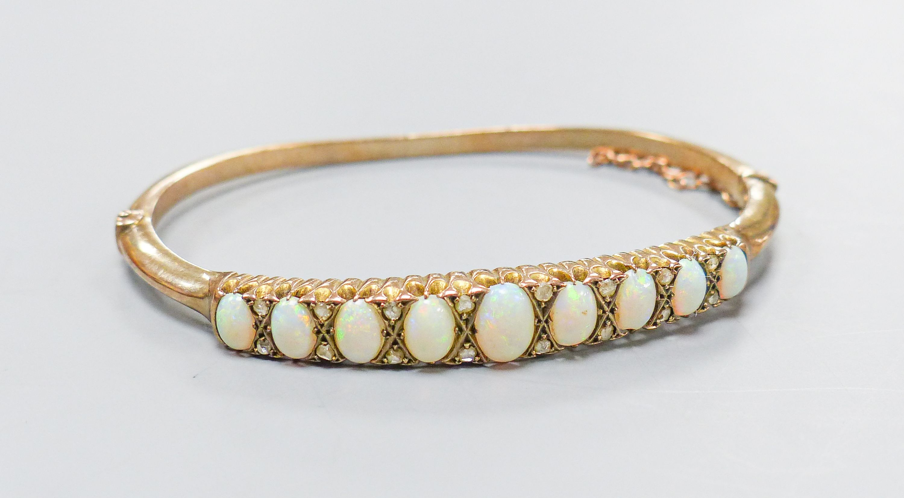 An early 20th century gold plated graduated nine stone white opal set hinged bracelet, with diamond chip spacers, in fitted box.